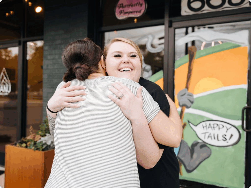 Two women embrace in a warm hug outside a storefront. The woman facing the camera has blonde hair and wears a black top, smiling broadly while hugging a woman with dark hair pulled back in a bun who wears a white and gray striped shirt. Behind them is a colorful storefront window with green and orange graphics and text including "HAPPY TAILS!" and what appears to be a pink sign reading "Recently Reopened" in the upper portion.