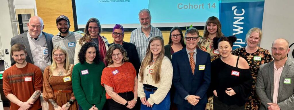 Group photo of the 14th ScaleUp WNC Cohort, approximately 17 participants posing together in two rows in front of a projection screen displaying 'mountainbizworks.org' and 'Cohort 14.' A blue ScaleUp WNC banner is visible on the right side. Many participants are wearing name tags. The group is diverse in age and appearance, smiling at the camera in what appears to be an indoor event or classroom setting.