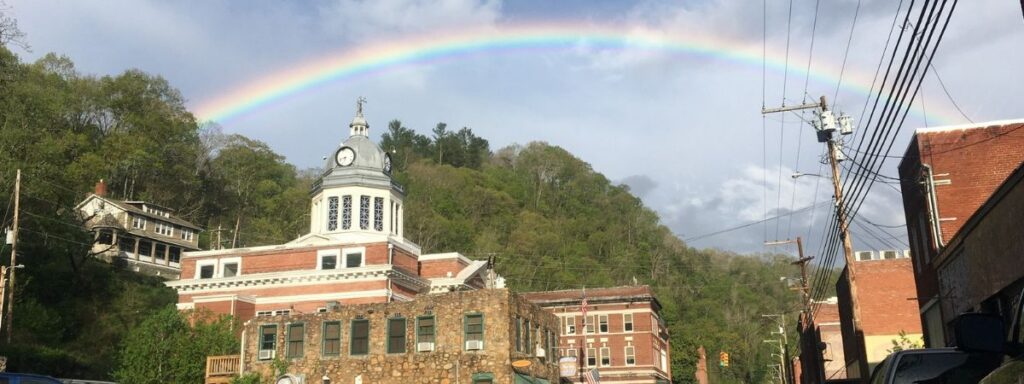 A full rainbow arches over the historic Madison County Courthouse and downtown Marshall, North Carolina, with tree-covered hills in the background.