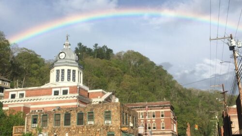 A full rainbow arches over the historic Madison County Courthouse and downtown Marshall, North Carolina, with tree-covered hills in the background.