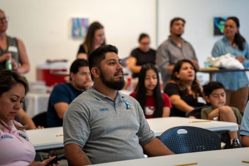 Ismael, a staff member from Rockhopper, sits attentively at a table during a community meeting or presentation. He wears a gray Rockhopper polo shirt. Several other adults and children are seated and standing in the background of the well-lit room.